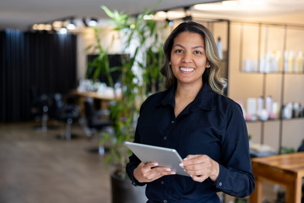 cosmetologist standing in front of hair salon smiling and holding tablet